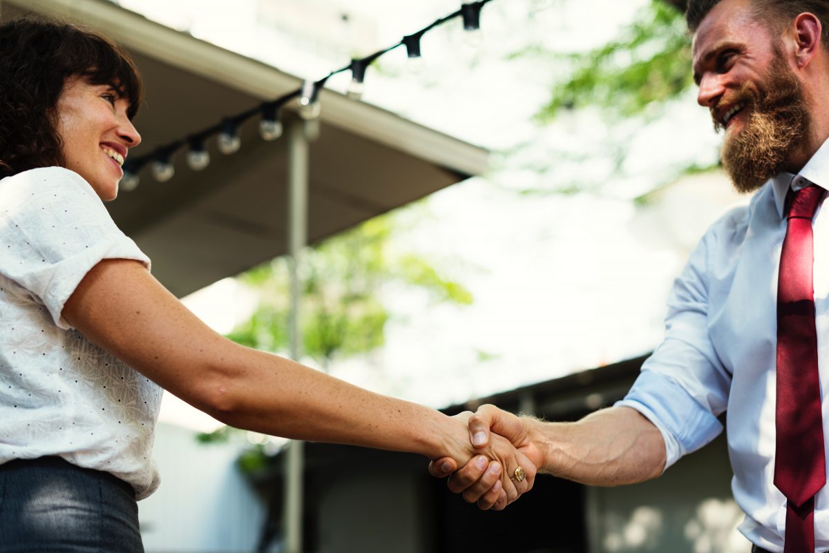 Eine Frau und ein Mann in Business-Kleidung schütteln sich die Hand und lächeln einander an.