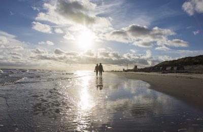 Ein Pärchen geht Barfuß am Meeresufer spazieren und laufen der Sonne entgegen.