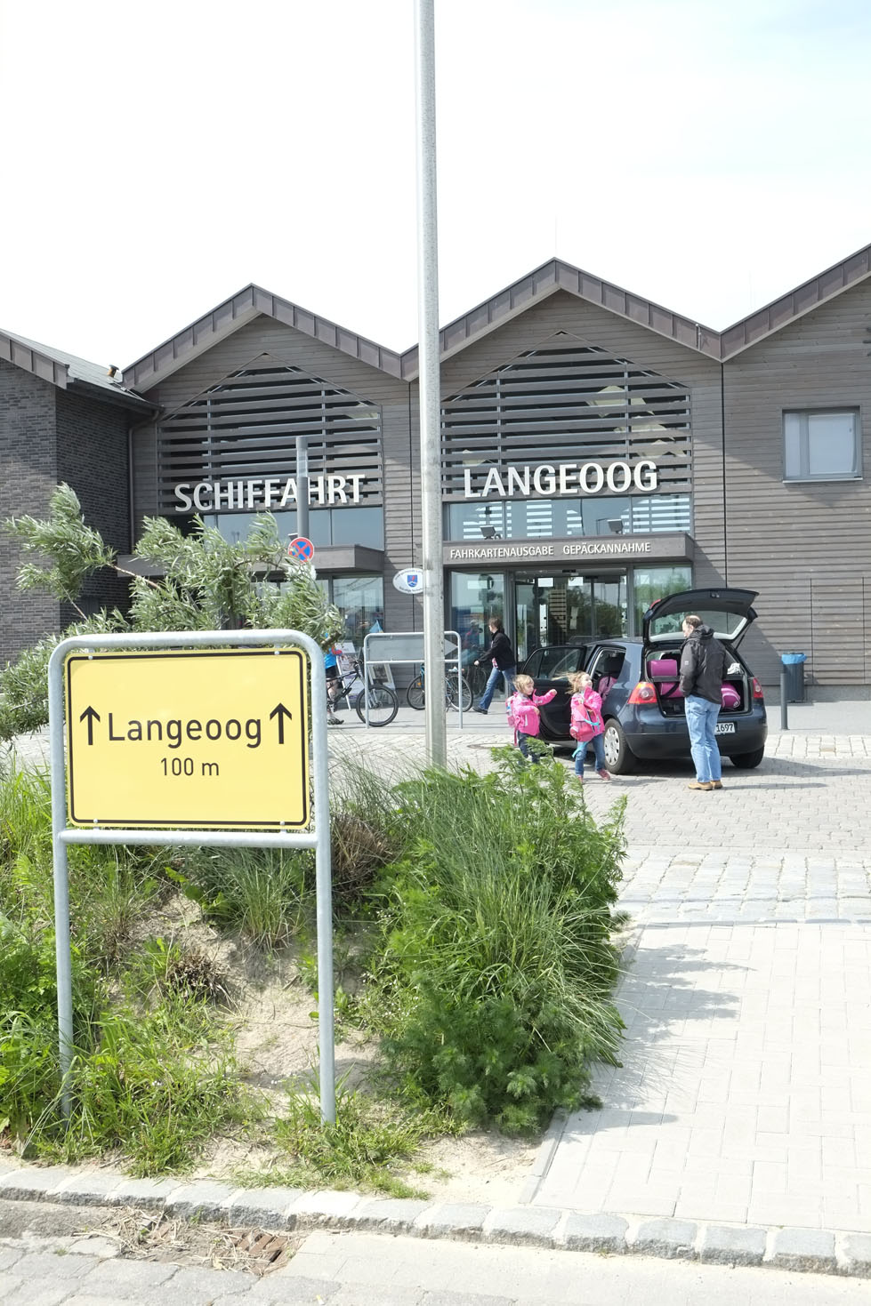 Das Gebäude der Schiffahrt Langeoog in der außenansicht. Vor dem Gebäude sind Menschen zu sehen, die Ihr Gepäck aus dem Kofferraum eines schwarzen Autos holen.