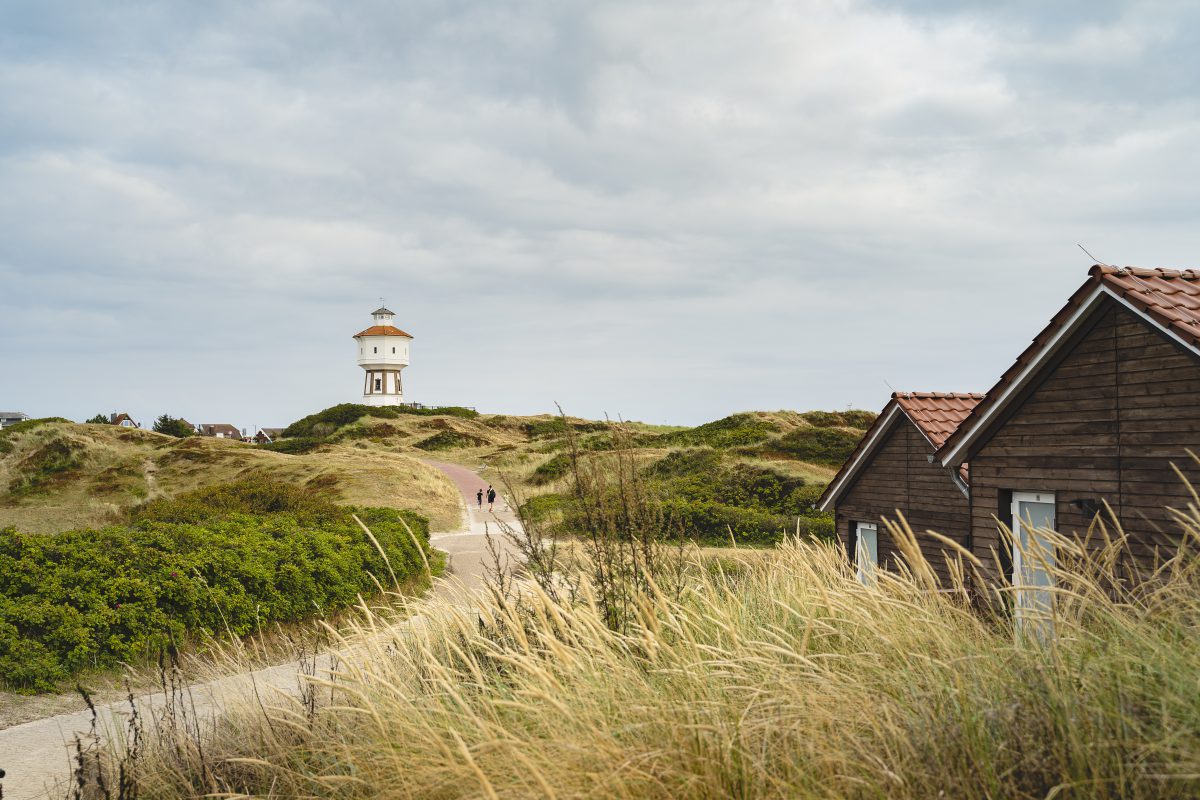 Zwei kleine Hütten in der Dünenlandschaft. Ein Weg mit Spaziergängern führt in Richtung Wasserturm.