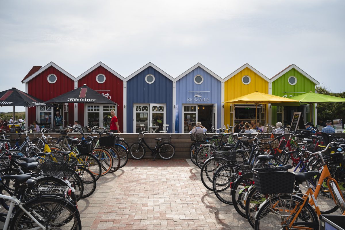 Eine kleine Reihe mit bunten Häusern, die verschiedene Geschäfte beherbergen. Die linken Häuser sind rot, die in der Mitte blau und rechts gibt es noch ein gelbes und ein grünes Häuschen. Hier sitzen viele Menschen draußen an Tischen mit Sonnenschirmen. Ein Zaun trennt diesen bereich von den Fahrradständern ab. Dort stehen sehr viele Fahrräder.