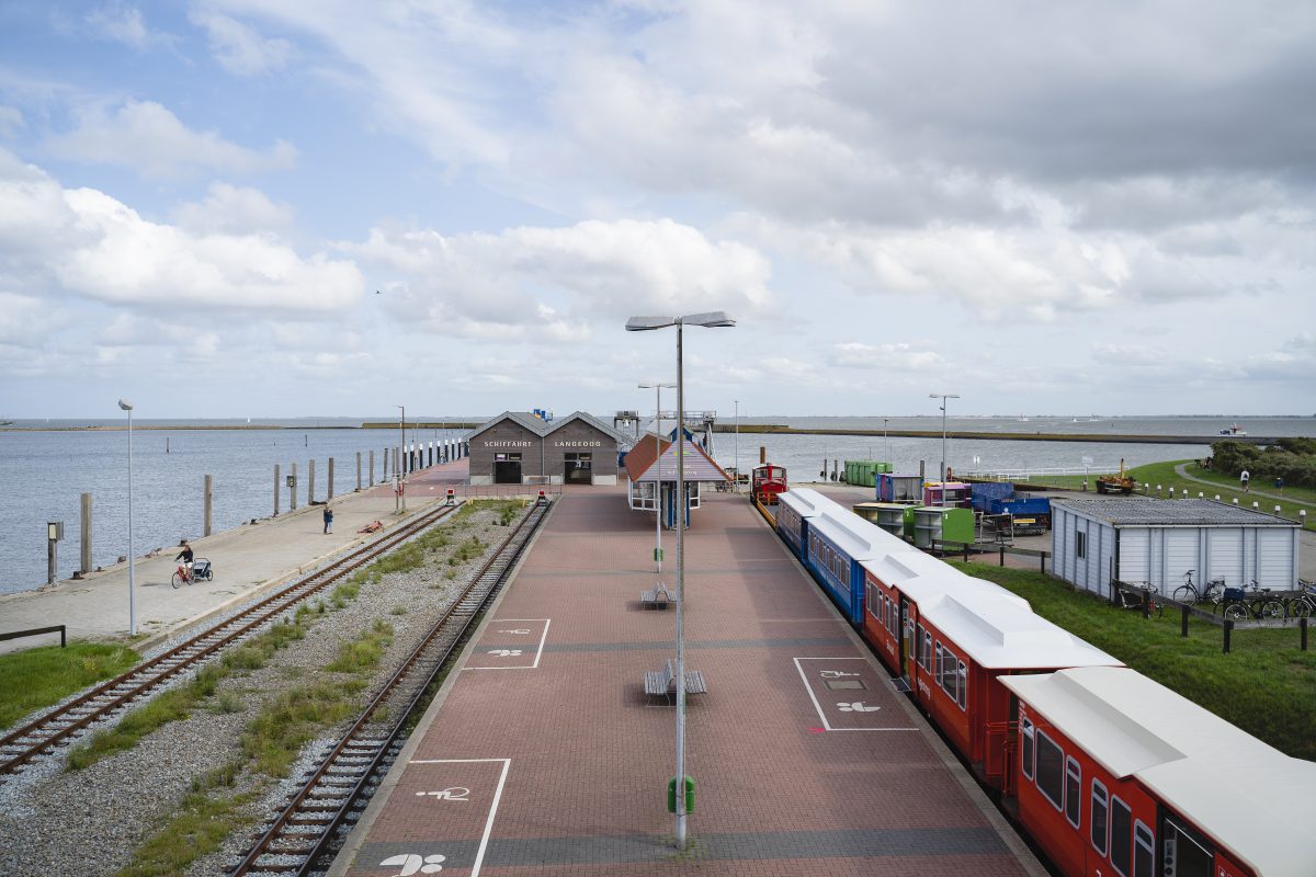 Der Anlegepunkt der Fähren aus der Froschperspektive. Man sieht den Durchgang der Schiffahrt Langeoog, die Inselbahn, Bahngleise, einen großen Bahnhofsplatz, eine grüne Landschaft und das Meer