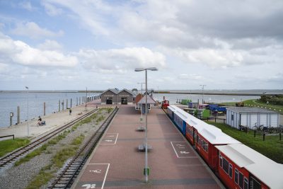 Der Anlegepunkt der Fähren aus der Froschperspektive. Man sieht den Durchgang der Schiffahrt Langeoog, die Inselbahn, Bahngleise, einen großen Bahnhofsplatz, eine grüne Landschaft und das Meer
